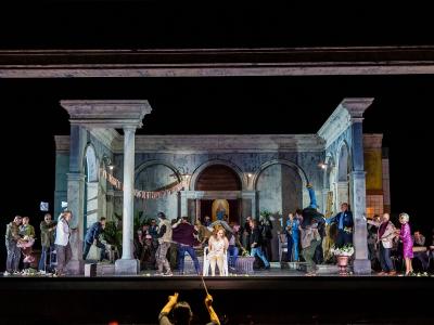 A theatrical stage performance at night featuring a set with classical columns and arches, with numerous performers on stage and a conductor's hands visible in the foreground.