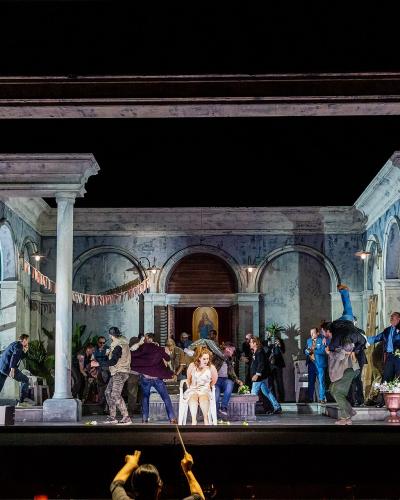 A theatrical stage performance at night featuring a set with classical columns and arches, with numerous performers on stage and a conductor's hands visible in the foreground.