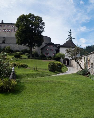 A stone castle with red and white window shutters sits atop a grassy hill, overlooking a courtyard with a stone wall and pathway.
