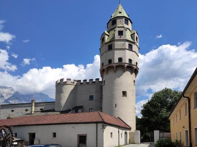 A tall, cylindrical stone tower with a distinctive green, multi-tiered conical roof and crenellated castle walls, set against a backdrop of mountains and a blue sky.