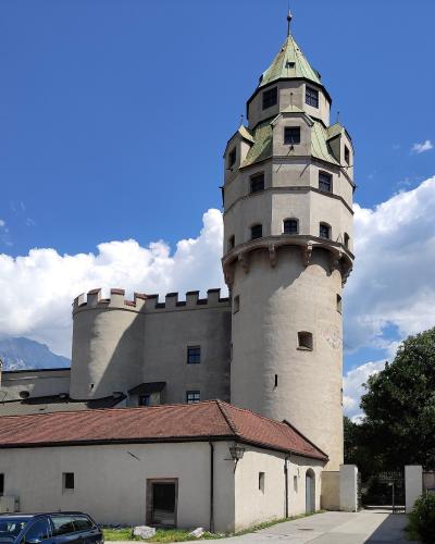 A tall, cylindrical stone tower with a distinctive green, multi-tiered conical roof and crenellated castle walls, set against a backdrop of mountains and a blue sky.