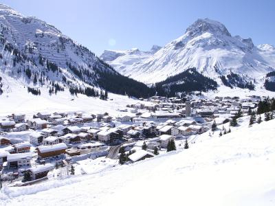 A snow-covered alpine village nestled in a valley surrounded by steep, snow-capped mountains and pine forests under a clear blue sky.