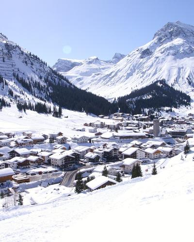 A snow-covered alpine village nestled in a valley surrounded by steep, snow-capped mountains and pine forests under a clear blue sky.