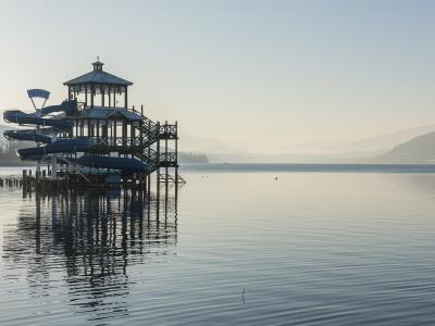 A wooden pier structure featuring a gazebo and multiple blue water slides extending over a calm lake, with mountains in the background.