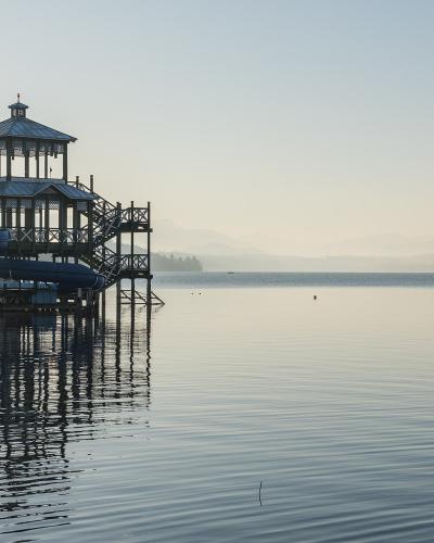 A wooden pier structure featuring a gazebo and multiple blue water slides extending over a calm lake, with mountains in the background.