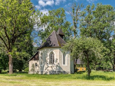 A small, white-walled chapel with a dark shingled roof and Gothic-style arched windows, situated in a grassy clearing surrounded by mature trees under a blue sky.