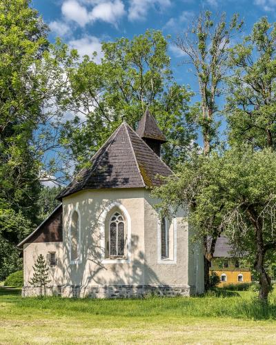 A small, white-walled chapel with a dark shingled roof and Gothic-style arched windows, situated in a grassy clearing surrounded by mature trees under a blue sky.