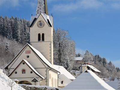 A white church with a tall, dark spire and a clock face, set against a snowy hillside with a small wooden structure in the foreground.