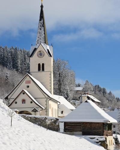 A white church with a tall, dark spire and a clock face, set against a snowy hillside with a small wooden structure in the foreground.