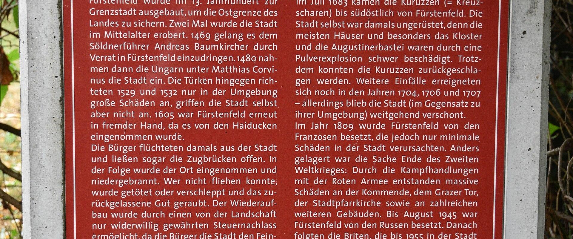 A red informational signpost for the "Festungsweg Fürstenfeld" trail, featuring text about the history of Fürstenfeld as a border city and a map of the fortification route with numbered stations.