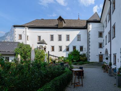 A white, multi-story historical building with grey stone quoins and a tiled roof, featuring a gravel courtyard with garden beds, closed umbrellas, and a mountain backdrop.