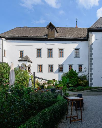 A white, multi-story historical building with grey stone quoins and a tiled roof, featuring a gravel courtyard with garden beds, closed umbrellas, and a mountain backdrop.
