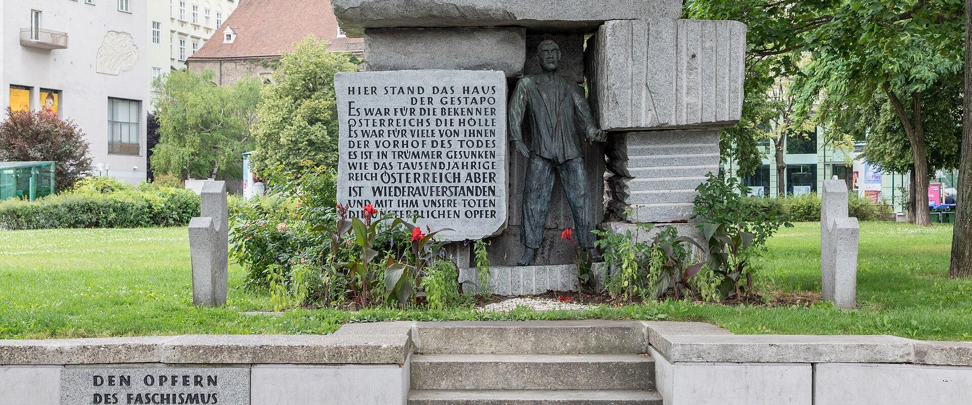 A stone memorial monument in a park-like setting in Vienna, featuring a bronze statue of a man, inscribed text including "NIEMALS VERGESSEN" and "DEN OPFERN DES FASCHISMUS BUNDESHAUPTSTADT WIEN", with a red triangle and yellow star symbol on the t