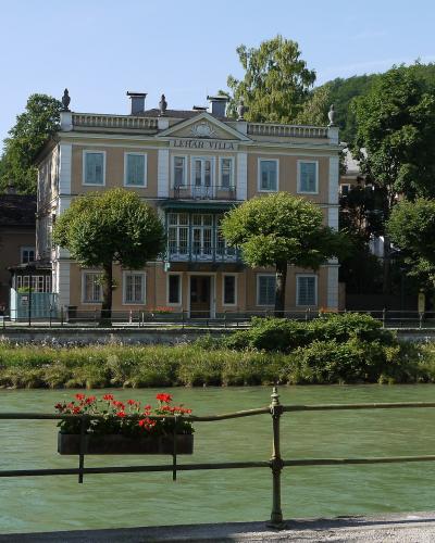 A large, classical-style yellow building with white trim stands on the bank of a green river, featuring prominent signage on its facade that reads "LEHAR VILLA".