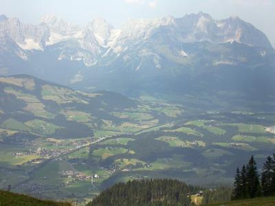 A high-angle landscape photograph showing a valley with scattered settlements and green fields, with a prominent, jagged, snow-dusted mountain range in the background.