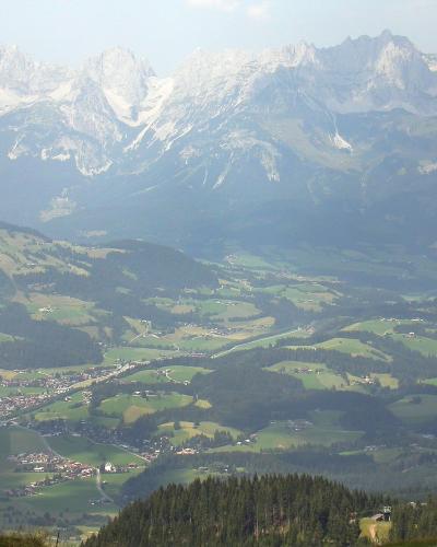 A high-angle landscape photograph showing a valley with scattered settlements and green fields, with a prominent, jagged, snow-dusted mountain range in the background.