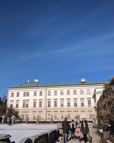 A large, cream-colored Baroque palace with a green roof stands behind a snow-covered formal garden, with visitors walking along a path next to a stone wall and trimmed hedges.