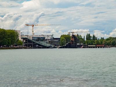 A large, temporary floating stage structure (seebühne) with a tall crane, situated on a lake, with a modern building and trees on the shore in the background.