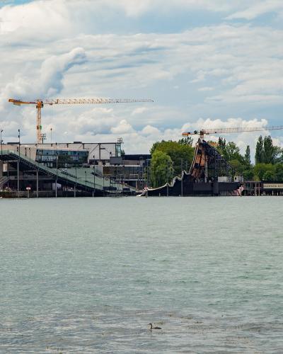 A large, temporary floating stage structure (seebühne) with a tall crane, situated on a lake, with a modern building and trees on the shore in the background.