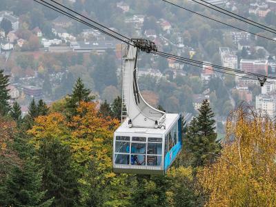 A close-up view of a white and blue cable car gondola suspended on cables, with a town and autumnal trees in the background.