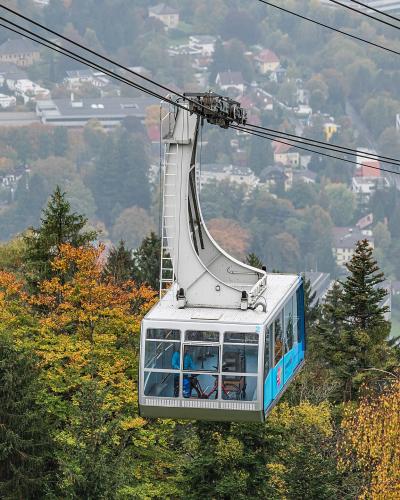 A close-up view of a white and blue cable car gondola suspended on cables, with a town and autumnal trees in the background.