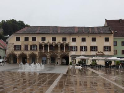 A large, historic building with a distinctive Gothic arcade and balcony facade, situated in a paved town square with a fountain and outdoor cafe seating under umbrellas.