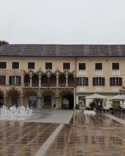 A large, historic building with a distinctive Gothic arcade and balcony facade, situated in a paved town square with a fountain and outdoor cafe seating under umbrellas.