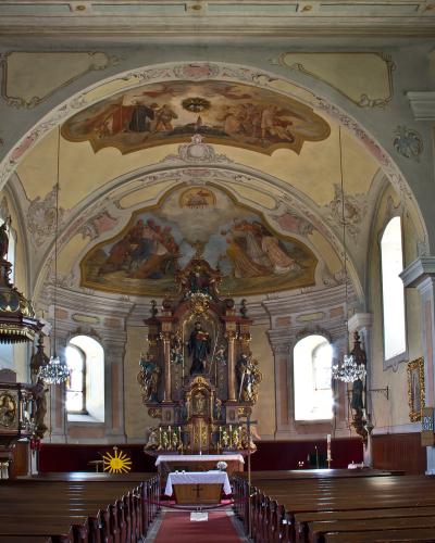 The image shows a large, ornate wooden altarpiece with multiple gilded statues of saints, situated inside a church with painted vaulted ceilings and frescoes on the walls.