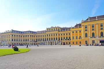 A wide-angle photograph of a large, yellow Baroque palace with a gravel courtyard in the foreground, green lawn to the left, and tourists walking around under a clear blue sky.