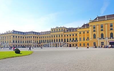 A wide-angle photograph of a large, yellow Baroque palace with a gravel courtyard in the foreground, green lawn to the left, and tourists walking around under a clear blue sky.