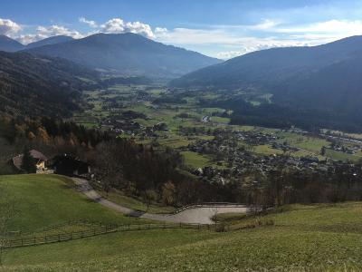 A high-angle landscape view of a wide, green valley floor with scattered houses and a winding road, framed by large forested mountains under a blue sky with clouds.