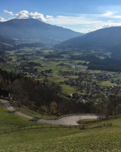 A high-angle landscape view of a wide, green valley floor with scattered houses and a winding road, framed by large forested mountains under a blue sky with clouds.