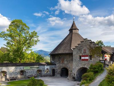 A stone castle gatehouse with a steep wooden roof and Austrian flags, set against a backdrop of green hills and mountains under a blue sky.