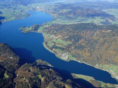 An aerial photograph showing a large, elongated alpine lake surrounded by forested hills and scattered settlements, with a prominent peninsula jutting into the water from the right side.