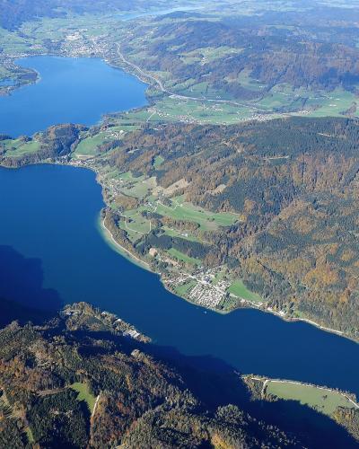 An aerial photograph showing a large, elongated alpine lake surrounded by forested hills and scattered settlements, with a prominent peninsula jutting into the water from the right side.