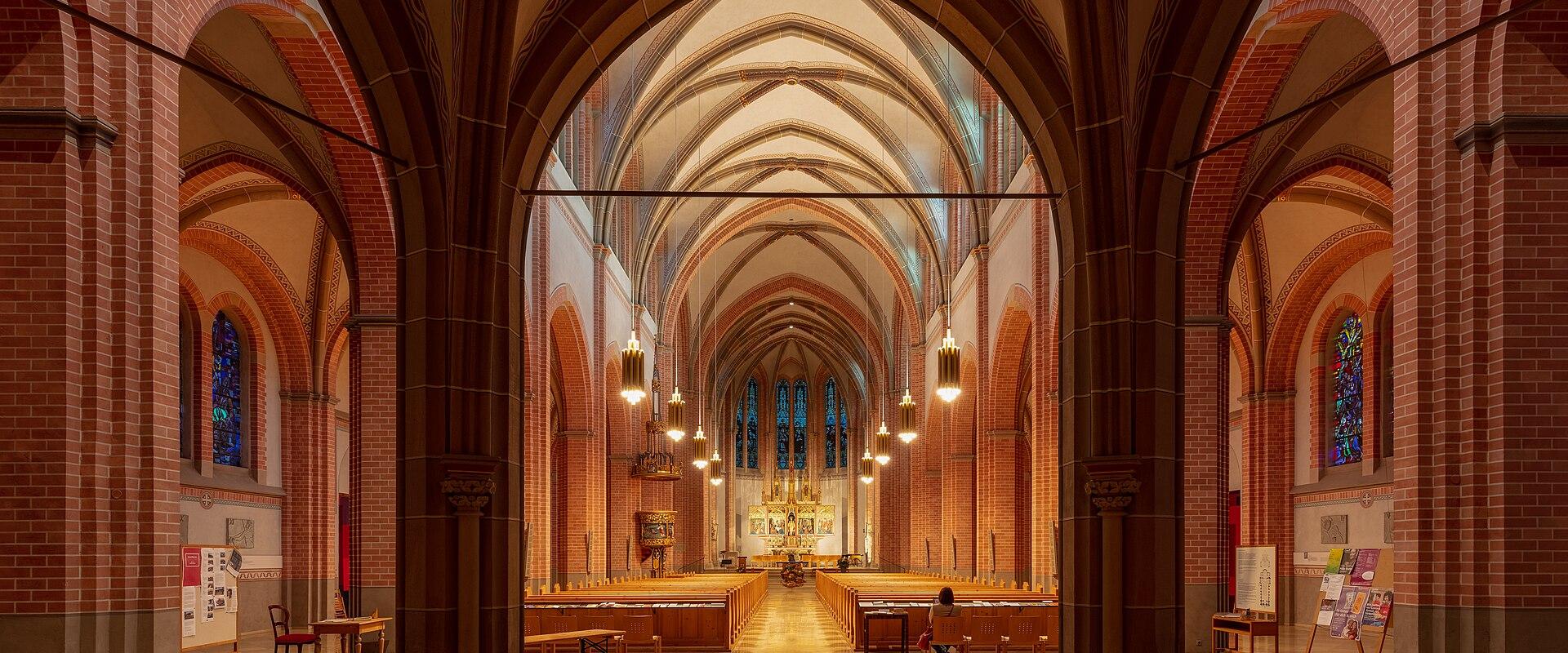 The interior of a large brick church with pointed arches, ribbed vaults, stained glass windows, and rows of wooden pews leading to an altar.