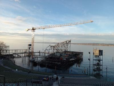 A view from the audience seating area of an open-air theater looking out onto a large floating stage structure under construction on a lake, featuring a tall yellow crane and scaffolding.