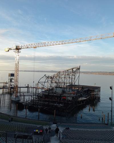 A view from the audience seating area of an open-air theater looking out onto a large floating stage structure under construction on a lake, featuring a tall yellow crane and scaffolding.