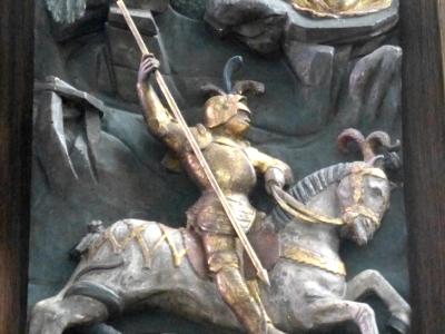 A close-up photograph of a painted wooden relief sculpture depicting the legend of Saint George slaying the dragon, with a princess observing from a tower in the background.