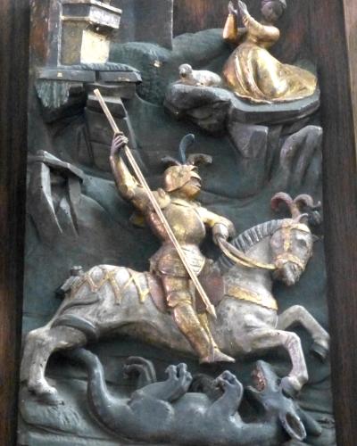 A close-up photograph of a painted wooden relief sculpture depicting the legend of Saint George slaying the dragon, with a princess observing from a tower in the background.