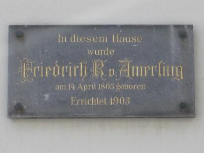 A close-up photograph of a grey commemorative plaque mounted on a light-colored wall, featuring gold lettering that reads "In diesem Hause wurde Friedrich R. v. Amerling am 14. April 1803 geboren Errichtet 1903".