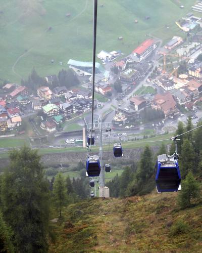 A high-angle view from a cable car looking down at the town of St. Anton am Arlberg, showing the village center, buildings, and surrounding green hills.