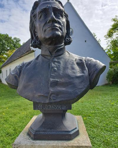 A bronze bust of Franz Liszt on a stone pedestal, with the inscription "F. LISZT" visible on the base, set outdoors on a grassy lawn with a white building featuring a steep gable roof in the background.