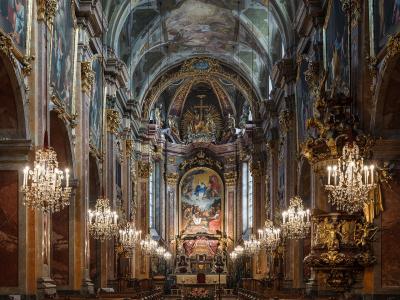 The interior of a grand Baroque cathedral nave looking towards the high altar, featuring ornate gold decorations, large crystal chandeliers, painted ceiling frescoes, and rows of wooden pews.