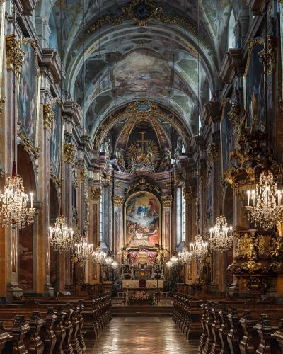 The interior of a grand Baroque cathedral nave looking towards the high altar, featuring ornate gold decorations, large crystal chandeliers, painted ceiling frescoes, and rows of wooden pews.
