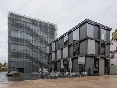 The image shows the Kunsthaus Bregenz, a modern art museum featuring a distinctive glass cube structure and an adjacent dark grey building with an outdoor terrace.