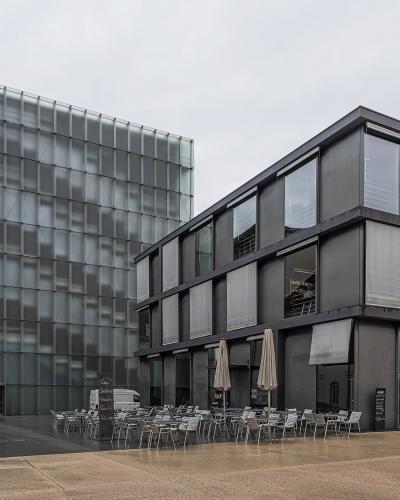 The image shows the Kunsthaus Bregenz, a modern art museum featuring a distinctive glass cube structure and an adjacent dark grey building with an outdoor terrace.