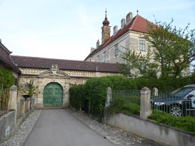 A stone gatehouse with a large green double door and a coat of arms above the archway, leading to a courtyard with a larger building featuring a tower and red roof in the background.