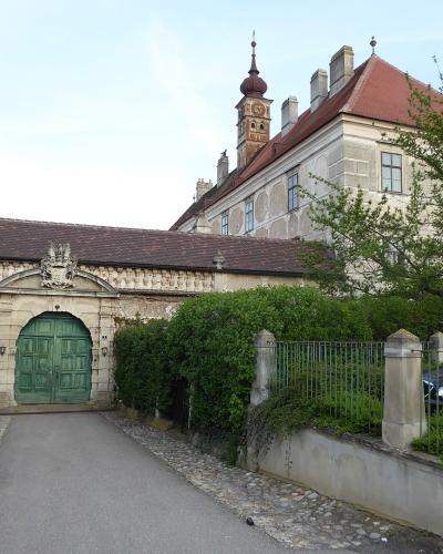 A stone gatehouse with a large green double door and a coat of arms above the archway, leading to a courtyard with a larger building featuring a tower and red roof in the background.