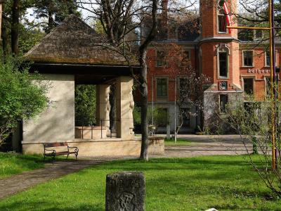 A red brick building with a corner tower and a stone gazebo in a park-like setting, featuring a visible "Rathaus" sign on the building facade.
